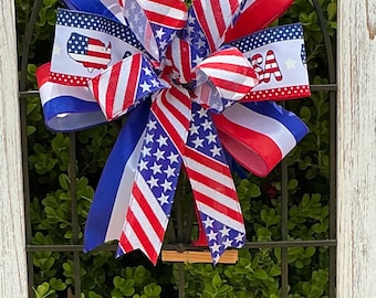 Op de afbeelding: Een patriottische strik met rood, wit en blauw lint. De strik bevat linten met de USA-kaart en sterpatronen. De strik is bevestigd aan een decoratief metalen frame, perfect voor Onafhankelijkheidsdag of andere patriottische vieringen.