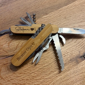 Könnte beinhalten: Ein Multitool mit Bambusgriff und verschiedenen Edelstahlwerkzeugen, darunter ein Messer, eine Säge und ein Korkenzieher. Der Griff ist mit dem Namen "Stefan" und einem Herzsymbol graviert. Der Text "Bester Opa" ist ebenfalls graviert.