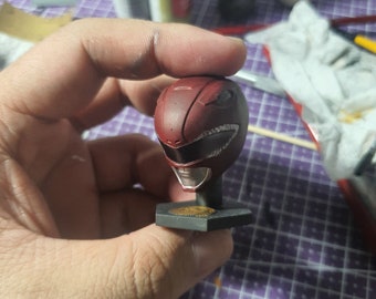 May include: A red plastic Power Ranger helmet with a white and black design. The helmet is on a black stand with a gold coin.