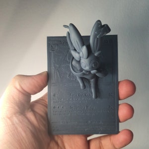 Op de afbeelding: Een grijze 3D-geprinte Pokémon-kaart met Sylveon, een Fee-type Pokémon. De kaart heeft de tekst "Time Out Kick" en "Symphony Whip" met overeenkomende aanvalswaarden van 30 en 70+. De kaart is bedrukt met een textuur.
