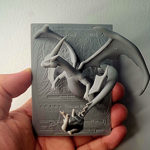 Könnte beinhalten: Ein 3D-gedrucktes Modell einer Charizard-Pokémon-Karte. Die Karte zeigt einen grauen und weißen Charizard mit roten Flügeln und dem Text "Charizard 220 KP" und "Glühender Sturz 100".