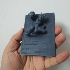 May include: A black and gray Pokemon card with a 3D printed figure of a black and gray Pokemon on top. The card features the text "Psyshock" and "70".