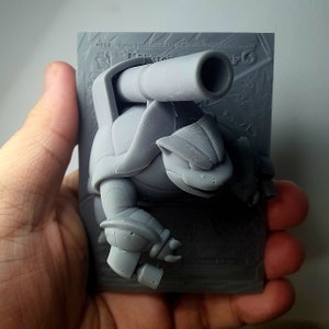 May include: A gray 3D printed figurine of a turtle-like creature with a cannon on its back. The figurine is on a gray card with the text "Mega:  Train your Pokemon to battle!  CPG".