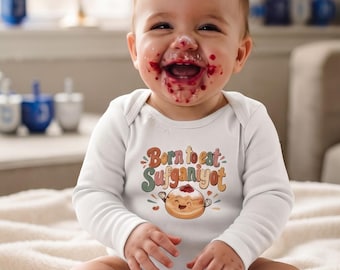 May include: A white long-sleeved baby bodysuit featuring a graphic of a jelly-filled donut with the text Born to eat Sufganiyot in colorful retro-style lettering. The garment has a round neckline and long sleeves, made of a plain white fabric.