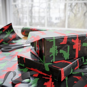Puede incluir: Dos cajas de regalo envueltas con papel de regalo negro con siluetas de armas rojas y verdes. También se ven un rollo de papel de regalo a juego, tijeras y una cinta negra, lo que sugiere un tema de envoltorio de regalo.