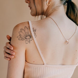 May include: A woman with a delicate flower tattoo on her shoulder. The tattoo is a simple line drawing of a daisy.