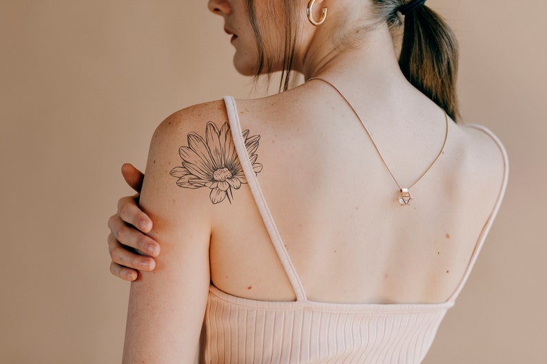 May include: A woman with a delicate flower tattoo on her shoulder. The tattoo is a simple line drawing of a daisy.