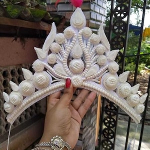 Op de afbeelding: Een witte kroon gemaakt van kleine, ronde, witte stukjes materiaal. De kroon is versierd met een rode veer bovenop.