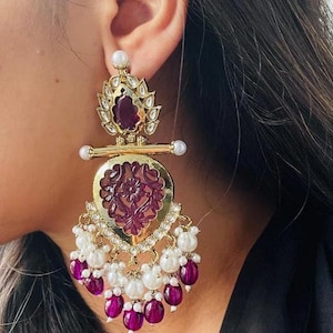 Puede incluir: Pendientes de oro con un diseño morado y blanco. Los pendientes presentan una piedra grande de color morado tallada con una piedra morada más pequeña en la parte superior. Los pendientes están adornados con perlas blancas y cuentas moradas.