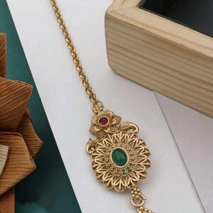 May include: A gold-tone maang tikka with a green and red stone design. The maang tikka has a chain with a hook at the top and a teardrop-shaped pendant with a red stone at the bottom.