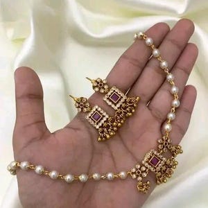 Może przedstawiać: Zestaw złotych naszyjnika i kolczyków z fioletowym kamieniem i białymi perłami. Naszyjnik to delikatny łańcuszek z zawieszką w kształcie kwadratu z fioletowym kamieniem w środku. Kolczyki to kolczyki wiszące o podobnym kwadratowym wzorze.