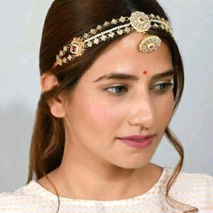 May include: A gold and white headpiece with a dangling red and white stone. The headpiece is worn on a woman's head and has a pearl and rhinestone design.