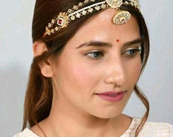 May include: A gold and white headpiece with a dangling red and white stone. The headpiece is worn on a woman's head and has a pearl and rhinestone design.
