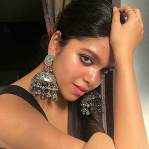 May include: A woman wearing large silver jhumka earrings with intricate designs and dangling charms. The earrings are a traditional Indian style.