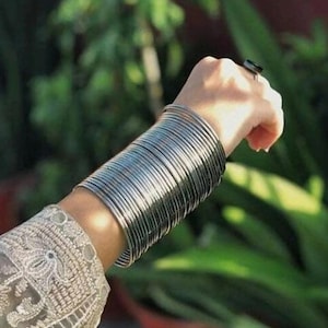 Op de afbeelding: Een stapel zilveren armbanden siert een pols en reflecteert licht. De dunne, metalen armbanden creëren een gelaagd effect. De mouw van een lichtgekleurd, geborduurd kledingstuk is zichtbaar.