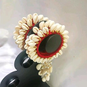 May include: A pair of red velvet bangles adorned with white seashells and gold beads. The bangles are displayed on a black stand with a decorative design.