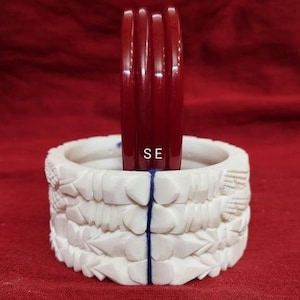 May include: A white carved decorative holder with three dark red bangles. The holder has a floral design and the letters "SE" are visible on the bangles. The background is a deep red.