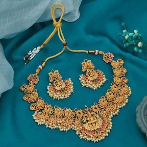 Può includere: Un set di collana e orecchini in oro con intricati disegni floreali, pietre preziose rosse e verdi e accenti di perle. La collana presenta un ciondolo centrale con una figura di divinità.