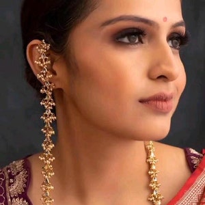 Puede incluir: Un par de pendientes largos de oro con detalles de perlas. Los pendientes están hechos de pequeños diseños intrincados y los lleva una persona con el pelo oscuro y maquillaje.