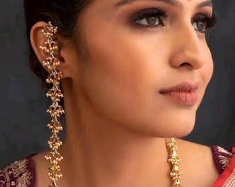 May include: A pair of long, gold earrings with pearl accents. The earrings are made of small, intricate designs and are worn by a person with dark hair and makeup.