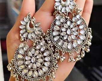 May include: A pair of silver earrings with a peacock design and a round, circular, crystal-like embellishment. The earrings have a dangling design with small beads.