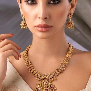 Peut inclure: Un collier en or avec un grand pendentif représentant une divinité. Le pendentif est entouré de petites perles dorées. Le collier est porté par une personne aux cheveux foncés et au maquillage.