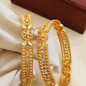 May include: Two gold bangles featuring elaborate floral patterns. The bangles are crafted from thick, weighty metal and have a polished finish.