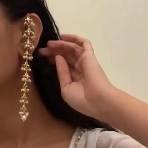 Könnte beinhalten: Ein langer, goldfarbener Ohrring mit weißen Perlenakzenten. Der Ohrring ist so konzipiert, dass er am oberen Ohr getragen wird, und verfügt über ein kaskadierendes Design aus kleinen, perlenartigen Perlen.