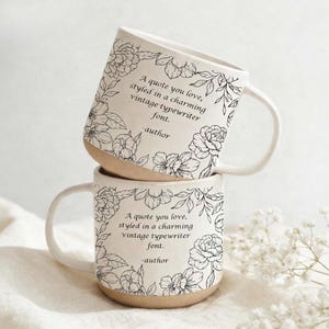 May include: Two white ceramic mugs stacked on top of each other. Each mug has a floral design with the text "A quote you love, styled in a charming vintage typewriter font. -author". The bottom of the mugs are tan.