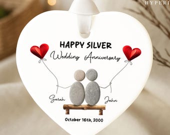 May include: White heart-shaped ornament with a ribbon for hanging. The ornament features the text "Happy Silver Wedding Anniversary" and a depiction of two pebble figures holding red heart balloons. Names "Sarah" and "John" are written below the figures, along with the date "October 16th, 2000."