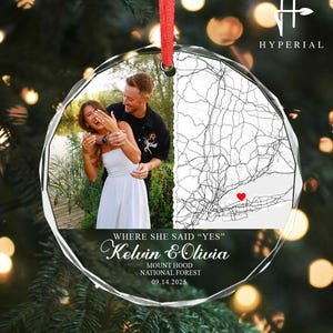 May include: A clear, round Christmas ornament with a red ribbon. The ornament features a photo of a couple and a map with a red heart. Text on the ornament reads "WHERE SHE SAID YES" and names "Kelvin & Olivia". The date 09.14.2025 is also visible.