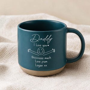 May include: A teal ceramic mug with a tan base. The mug has a curved handle and features the text "Daddy I Love you" and a drawing of a smiley face with outstretched arms. The text "thiiiiisssss much Love from Logan xx" is also on the mug.