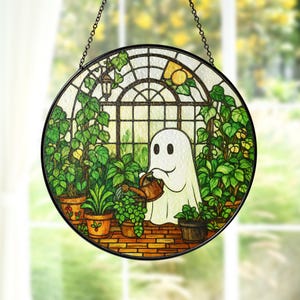 May include: Round stained glass window art featuring a friendly ghost watering plants in a greenhouse. The design includes a brick floor, potted plants, and a window with a lemon and a lantern. The artwork is framed in black and hangs from a chain.