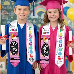 May include: Two children in graduation gowns, one blue and one pink, wearing sashes that say "Preschool" and "Class of 2026." Each child holds a diploma. The sashes are pink with a crayon and pencil design.