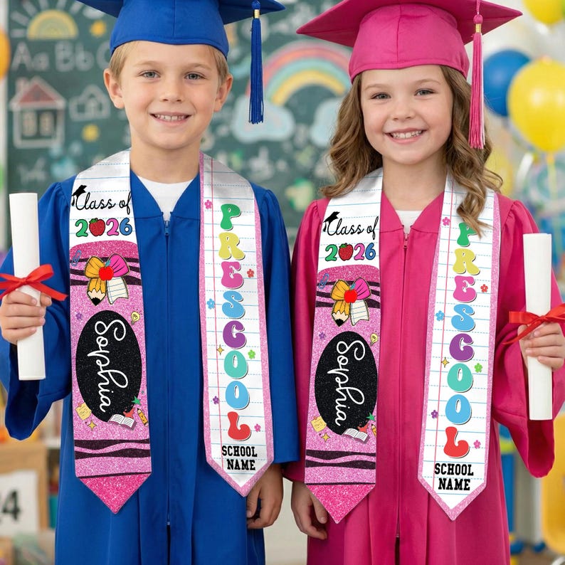 May include: Two children in graduation gowns, one blue and one pink, wearing sashes that say "Preschool" and "Class of 2026." Each child holds a diploma. The sashes are pink with a crayon and pencil design.
