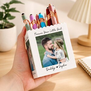 May include: A white pen holder with a photo of a father and daughter, and the text "Forever Your Little Girl" and "Daddy Sophia". The holder contains several pens in various colors, including green, purple, and pink.