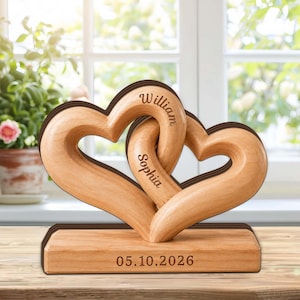 May include: A wooden sculpture featuring two interlocking hearts. The names "William" and "Sophia" are engraved on the hearts. The base displays the date "05.10.2026". The sculpture is made of light-coloured wood.