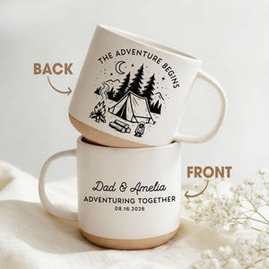 May include: Two white ceramic mugs stacked on top of each other. The top mug has a black camping scene with the text "THE ADVENTURE BEGINS". The bottom mug has the text "Dad & Amelia ADVENTURING TOGETHER 08.16.2026".