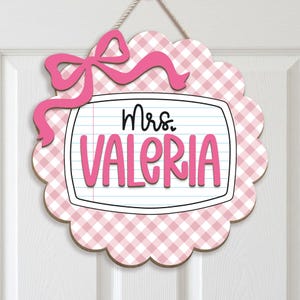 May include: A scalloped circular wall sign featuring a pink and white gingham pattern with a pink bow at the top. The centre displays a white rectangular panel resembling notebook paper with the black text Mrs. and the pink text VALERIA.