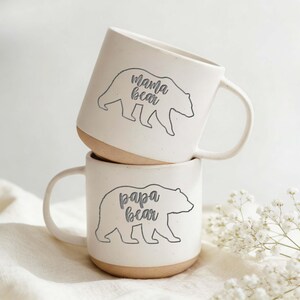 May include: Two stacked white ceramic mugs with a tan base. The top mug has "Mama Bear" and a bear outline, the bottom mug has "Papa Bear" and a bear outline. Both mugs have a curved handle.