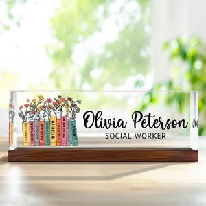 May include: A clear acrylic desk sign with a wooden base. The sign features the name "Olivia Peterson" and the title "SOCIAL WORKER" in black script. Colourful books with floral arrangements and inspirational words are on the left.