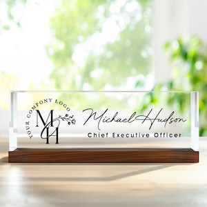 Peut inclure: Une plaque de bureau rectangulaire en acrylique transparent posée sur un socle en bois brun foncé. L'avant présente un texte imprimé en noir indiquant "YOUR COMPANY LOGO", les initiales "MH" avec un motif floral, "Michael Hudson" et "Chief Executive Officer".