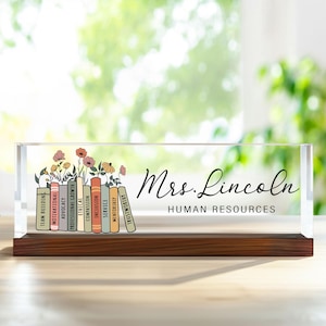 May include: A clear acrylic desk block with a wooden base. The block features a floral design above a row of colorful book spines with words like "Team Building" and "Motivational." The name "Mrs. Lincoln" and "Human Resources" are written in black script.