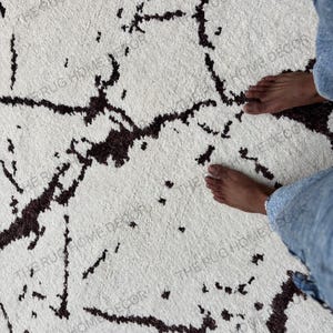 May include: A white rug with a dark brown, abstract pattern. The rug has a soft, textured appearance. The words "THE RUG HOME DECOR" are printed on the rug.