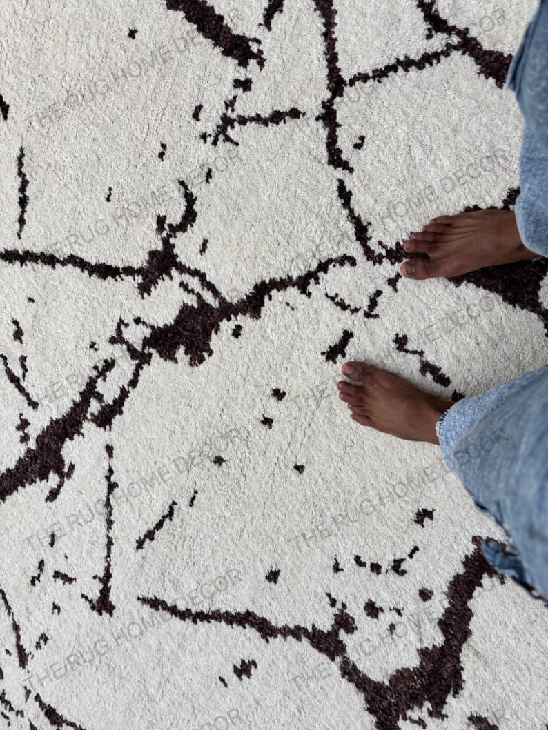 May include: A white rug with a dark brown, abstract pattern. The rug has a soft, textured appearance. The words "THE RUG HOME DECOR" are printed on the rug.