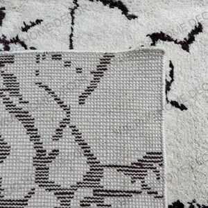 May include: Close-up of a beige rug with a geometric pattern in dark brown. The rug has a textured surface and a visible border. The words "HOME DECOR" are visible in the image.