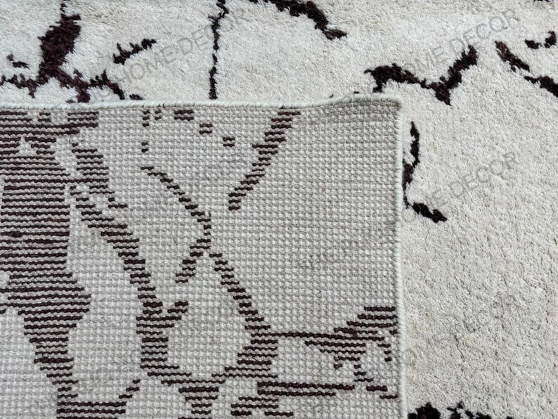 May include: Close-up of a beige rug with a geometric pattern in dark brown. The rug has a textured surface and a visible border. The words "HOME DECOR" are visible in the image.