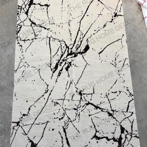 May include: Rectangular area rug with a white background and a black, marble-like pattern. The rug has a textured surface and features the text "THE RUG HOME DECOR" in multiple places.