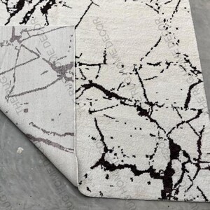 May include: A white rug with a black marble-like pattern. The rug is partially rolled up, revealing the underside with text that reads "RUG HOME DECOR". The design features irregular black lines and spots across the white surface, creating a modern aesthetic.