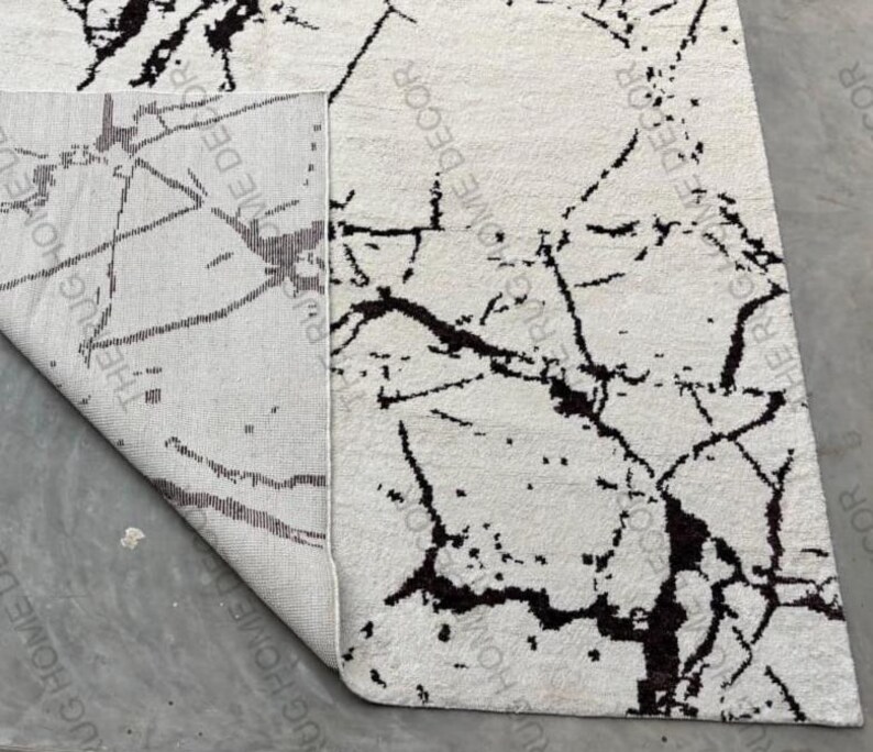 May include: A white rug with a black marble-like pattern. The rug is partially rolled up, revealing the underside with text that reads "RUG HOME DECOR". The design features irregular black lines and spots across the white surface, creating a modern aesthetic.